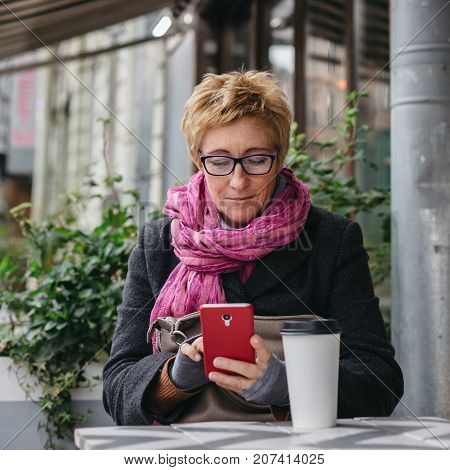 Smiling adult woman browsing smartphone and sitting with paper cup in outside cafe.