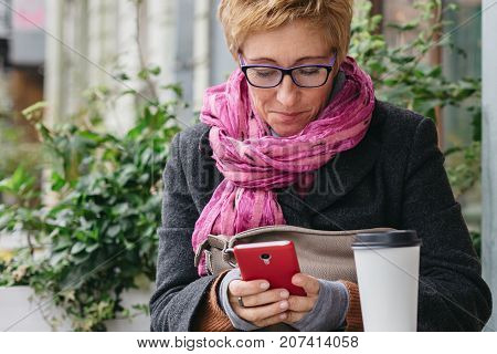 Smiling adult woman browsing smartphone and sitting with paper cup in outside cafe.