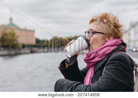 Side view of adult blond woman drinking coffee while standing at water on esplanade.