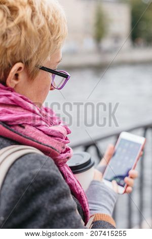 Adult woman in glasses standing with smartphone and coffee on waterfront.