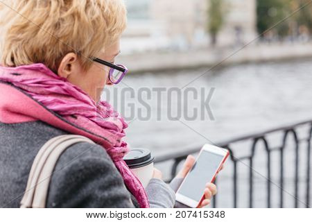 Adult woman in glasses standing with smartphone and coffee on waterfront.