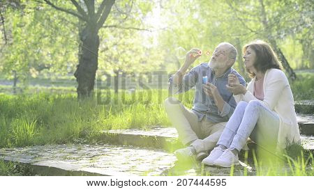 Beautiful senior couple in love outside in spring nature, sitting on stairs, blowing bubbles with bubble wand.