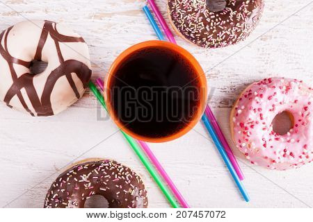 Overtop view of donuts, a glass of cola next to straws on white wooden background