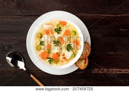 An overhead photo of a plate of chicken, vegetables, and noodles soup, shot from above on a dark rustic texture with slices of bread and a spoon, and a place for text