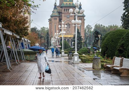 TIMISOARA ROMANIA - SEPTEMBER 21 2017: Old woman with an umbrella walking on the iconic Victory Square (Piata Victorei) the Metropolitan Cathedral (Catedral Mitropolitana) can be seen in the background