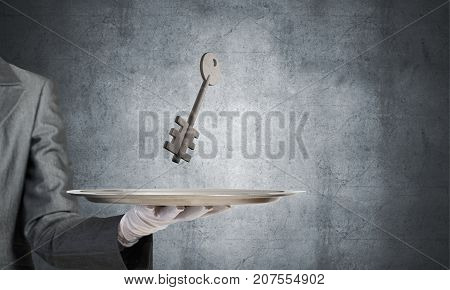 Closeup of waiter's hand in white glove presenting stone key symbol on metal tray with gray wall on background. 3D rendering.