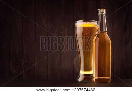 Transparent beer bottle and glass weizen with golden lager on dark brown wood board copy space mock up. Template for advertising design branding identity.