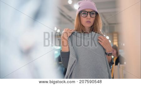 A young stylish hipster woman in fashionable glasses and a hat tries on new clothes in the store. Shopping