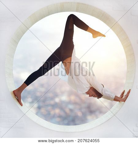 Beautiful sporty fit yogi woman practices yoga asana eka pada Adho Mukha Shvanasana One Leg Downward Dog pose in a round window with a view of the city at sunset.