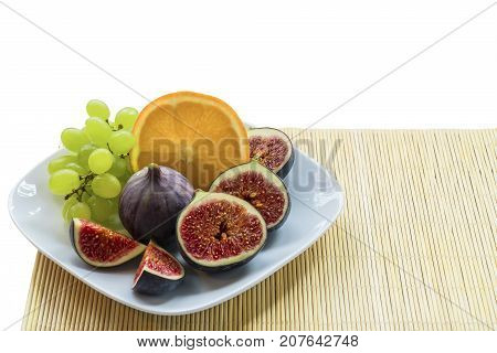 White plate with summer juicy sweet dietary exotic fruits of figs grapes orange on a bamboo napkin.