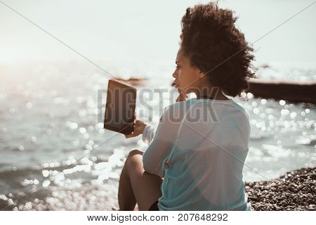 Rear view of attractive young Afro American female sitting on the beach of summer resort with ocean in defocused background and taking selfie using frontal camera of her digital tablet; summer day