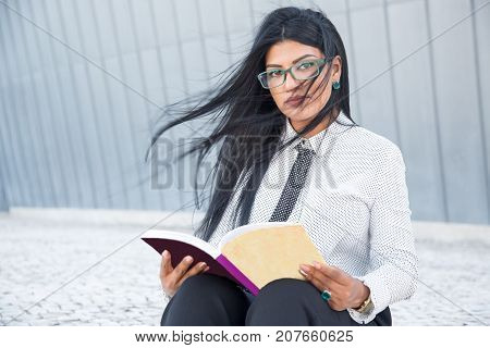 Confident smart woman reading business book and looking at camera. Serious beautiful businesswoman in eyeglasses interested in self-education. Student concept