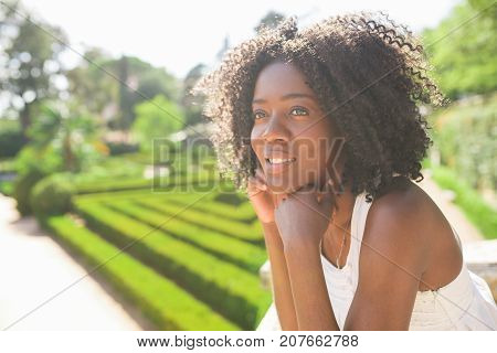 Closeup portrait of smiling young attractive African American woman standing and leaning on railing in park with blurred beautiful garden in background. Side view.