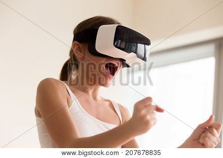 Young female gamer in wearable VR headset playing action 3d video game at home, excited teen holding wheel and screaming driving digital racing car in simulator application, virtual gaming concept