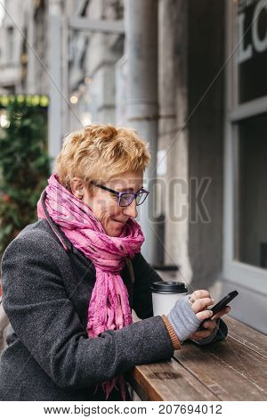 Adult blonde woman sitting with coffee and smartphone in outside cafe on street.