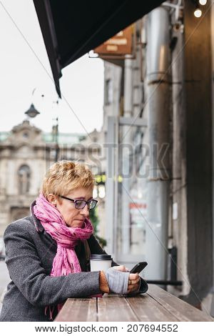 Adult blonde woman sitting with coffee and smartphone in outside cafe on street.