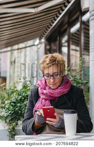 Smiling adult woman browsing smartphone and sitting with paper cup in outside cafe.