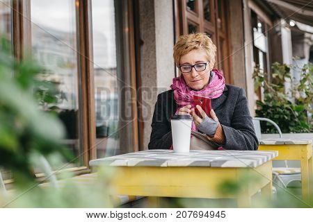 Adult blonde woman browsing smartphone and having drink from paper cup in outside cafe.