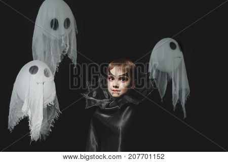 Vampire boy expressive portrait staring eyes, wearing a black cloak with high bat like collar looking at camera on black studio background, with flying white ghosts. Halloween vampire makeup.
