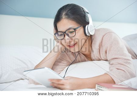 Happy Beautiful Asian woman listening to the music with headphone and tablet or mobile phone on her bed. Blue wall.