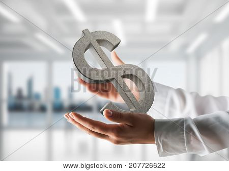 Closeup of business woman in white shirt keeping stone dollar sign in hands with office view on background. Mixed media.