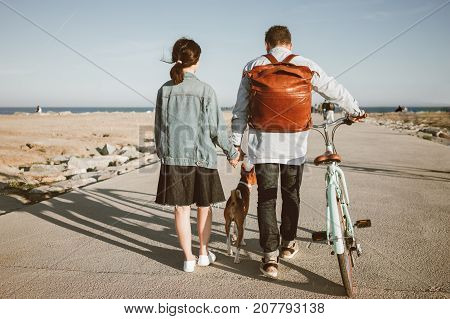 Girl in denim dress and jacket with man in jeans and blue shirt with leather brown backpack. Couple walks to sea on beach with basenji dog and holds hands guy leads girlfriend's mint bike. Overall view from back.