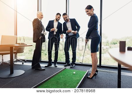 Three men watch as their boss plays mini-golf in her office. She is holding a golf club.