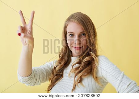 Picture of positive cheerful young female making peace sign looking at camera with happy friendly smile. Attractive woman posing in studio making V gesture which stands for victory. Body language