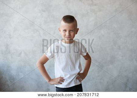 Studio picture of naughty mischievous Caucasian preschool boy dressed in casual white t-shirt holding hands on his waist and squinting eyes planning another trick showing his stubborn temper