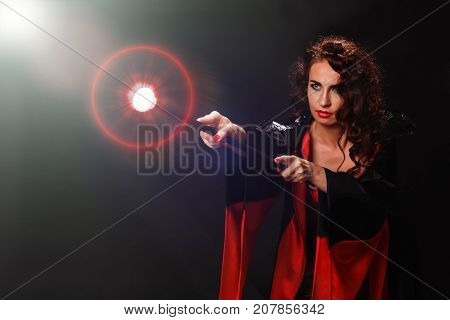 A young beautiful red-haired woman in the guise of a witch, a witch in a cloak of black and black fabric holds in her hand a magic staff that emits a bright light, a fireball, a rainbow