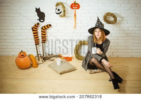 Halloween coffee or tea break. Woman in witch hat with cups. Girl sitting on wooden floor. Holiday celebration concept. Pumpkins stockings black cat wreaths mummy decorations on brick wall.