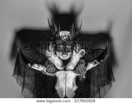 Gothic man in masquerade mask with feathers on his face long white hair and beard in black theater costume on background of big shadow holding cranium of horned animal