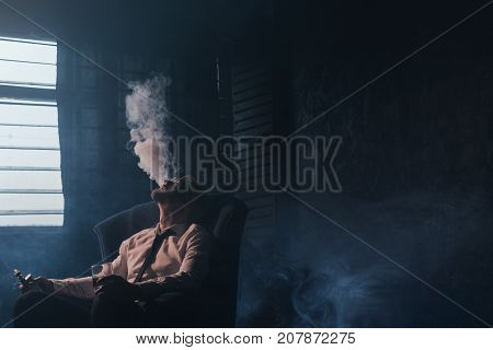 Businessman relaxing with vape and alcohol. Dark room with white atmospheric smoke background, rich people entertainment, private club for luxury life with free space