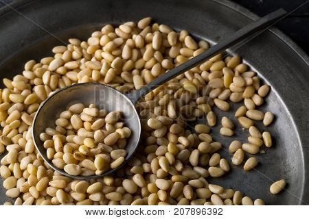 Heap of raw uncooked pine nuts in metal spoon on rustic plate over dark kitchen background