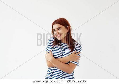 Cute girl with red colored hair crossing arms on her chest and hugging herself having joyful happy look standing isolated against white studio wall background with copy space for your text