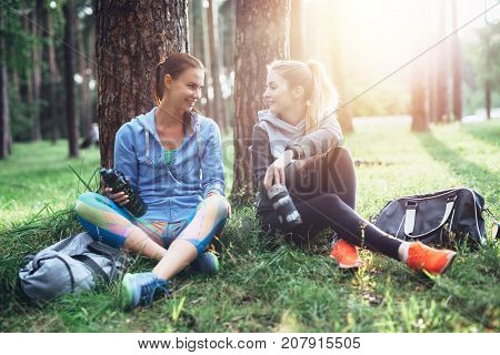 Two young women in sportswear sitting under trees in the forest drinking water, talking and resting after training outdoors.