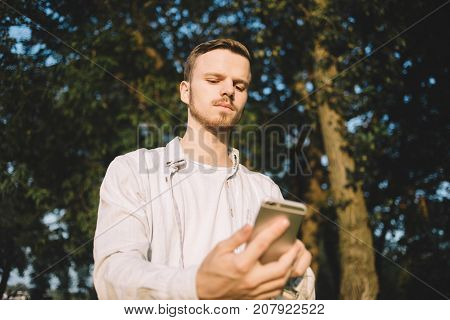 A picture of a young guy standing near green trees in a beautiful evening time. The young man is looking to the phone's screen with a serious and concentrated sight. Clse up. Cut view