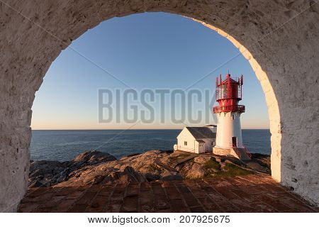 Lindesnes Fyr, historic lighthouse in Norway, seen through a rounded window in a nearby building. Red and white lighthouse on the edge of rocky sea coast in the southern most place in Norwegian mainland
