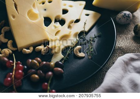 Cheese with holes lying on stylish black dish, decorated with berries, nuts and cashew. Snacks for dining and drinking wine in a good company. 