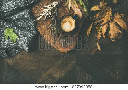 Autumn or Fall morning coffee concept. Flat-lay of arm knitted woolen grey jamper, wooden tray, mug of coffee and yellow fallen leaves over dark rustic wooden table background, top view, copy space