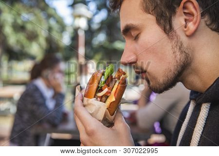 Hungry Young Man Enthusiastically Sniffing His Burger While Sitting At The Restauraunt Terrace. His 