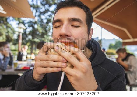 Young Man Is Greedily Eating His Burger While Sitting At The Restauraunt Terrace. His Eyes Are Close