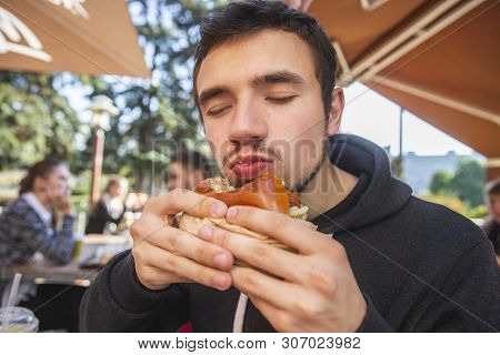 Satisfied Young Man Is Holding His Bitten Burger, His Eyes Are Closed. He Is Sitting At The Fastfood