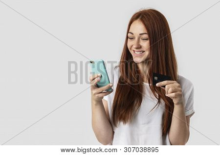 Young Red-haired Girl Isolated On Gray Background In White T-shirt. She Holds The Phone And Credit C