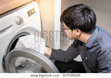 Asian Man Loading White Clothes Into Front Door Washing Machine In Laundry Room. Housework Or Chores