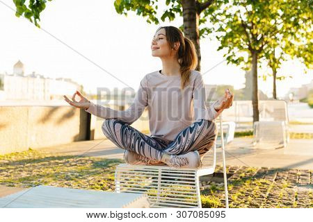 Cheerful young fitness woman relaxing outdoos, sitting on a bench, listening to music with earphones, meditating