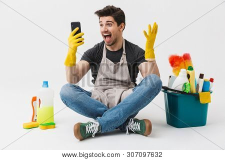 Image of a happy shocked young houseman househusband with cleansers on floor isolated over white wall background make winner gesture using phone.