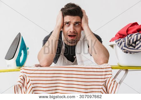 Image of a tired bored young houseman househusband with iron and clothes isolated over white wall background.
