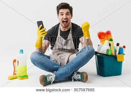 Image of a happy excited young houseman househusband with cleansers on floor isolated over white wall background make winner gesture using phone.