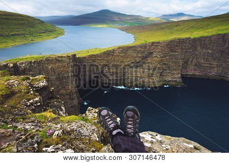 Legs Of Hiker Sitting On Top Of A Cliff With Views Over Cliff Traelanipan And Lake Sorvagsvatn Locat